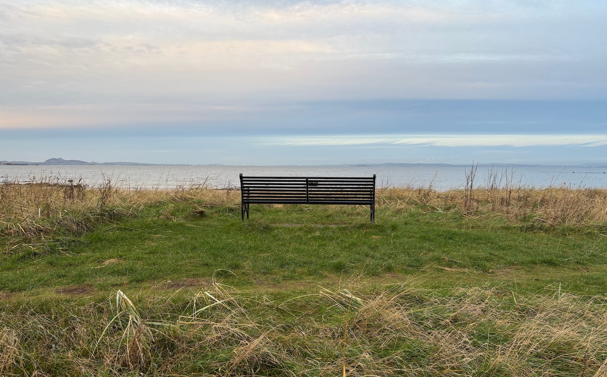 A black metal bench on sand dunes, overlooking the sea, with Arthur's Seat and the Fife coast in the distance